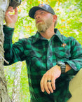 Man in green plaid wool shirt and cap holding a device against a tree in a forest