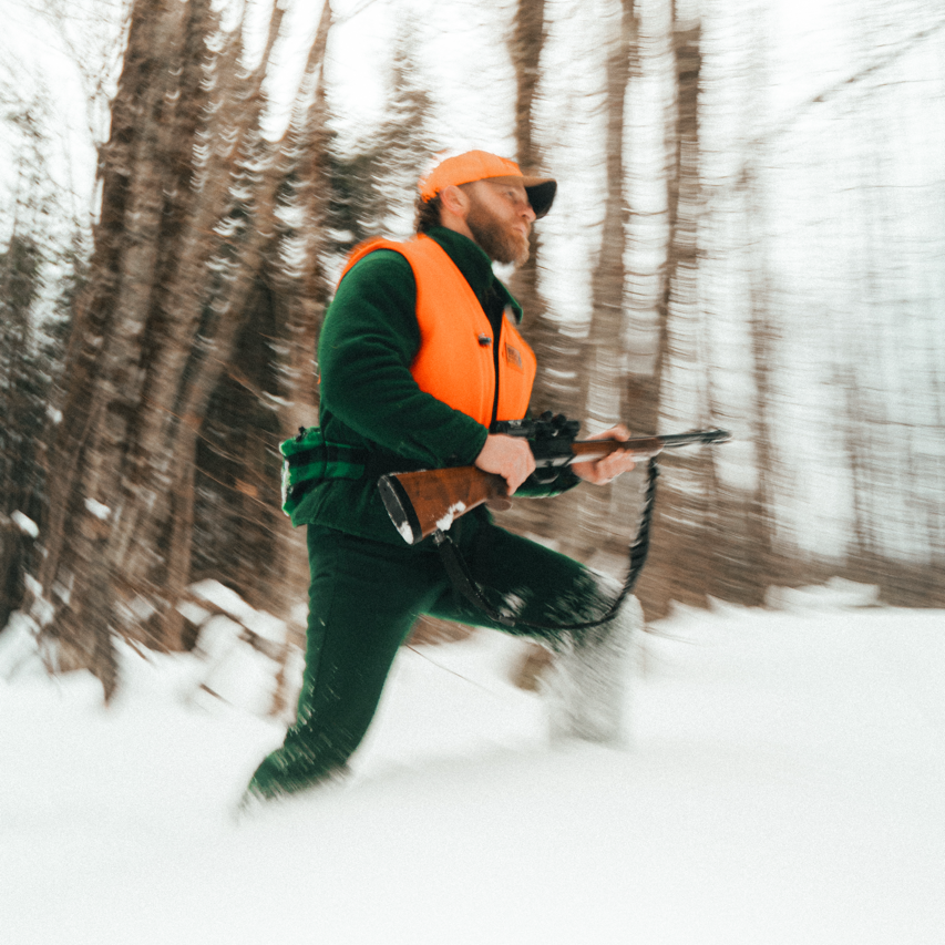 Hunter in cold weather wool hunting gear with a rifle walking through a snowy forest.