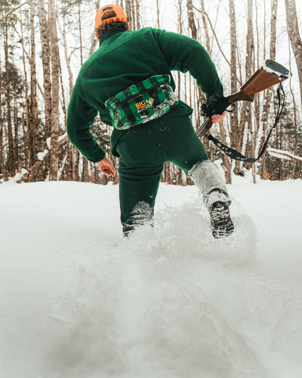 Person walking through deep snow carrying a rifle, wearing wool hunting gear  in a snowy forest setting